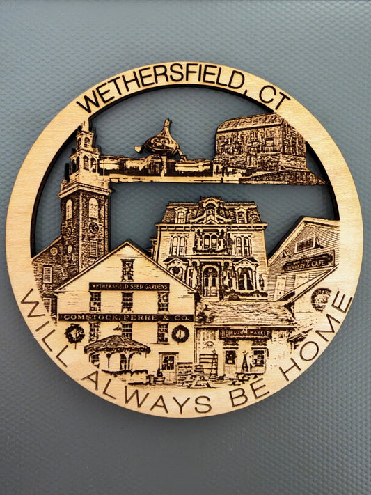 A circular laser-engraved wooden plaque featuring a scenic cutout illustration of Wethersfield, CT, with the text 'WETHERSFIELD, CT' at the top and 'I WILL ALWAYS BE HOME' along the bottom. The natural wood plaque is displayed on a textured gray background.